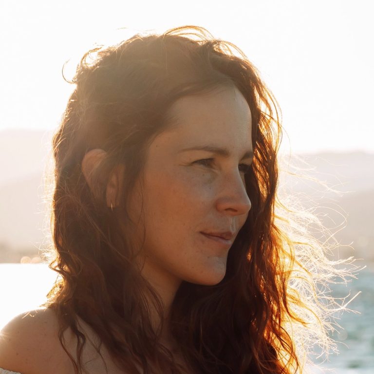 Femme au bord de l'eau, cheveux bouclés, regard tourné vers l'horizon ensoleillé.