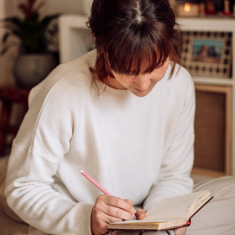 Femme écrivant dans un carnet avec un crayon, assise sur le sol.